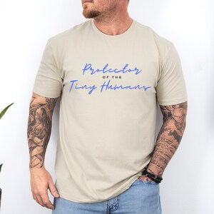 May include: A beige t-shirt with the text "protector of the tiny humans" in blue script.