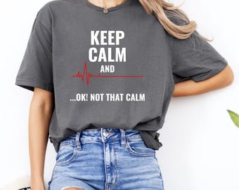 Puede incluir: Camiseta gris oscuro con el texto "KEEP CALM AND ...OK! NOT THAT CALM" en blanco y rojo. La camiseta presenta un gráfico de línea de latido cardíaco rojo. La modelo lleva vaqueros azules.