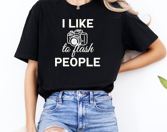 May include: Black t-shirt with the text "I LIKE to flash PEOPLE" in white, along with a camera graphic. The shirt is paired with blue jeans.