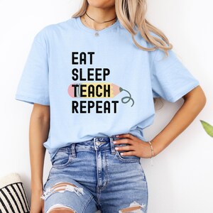 Puede incluir: Una camiseta azul claro con el texto "Eat Sleep Teach Repeat" y un gráfico de lápiz de dibujos animados.