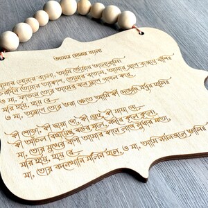 May include: A wooden plaque with a Bengali poem engraved on it. The poem is about love for one's homeland. The plaque is decorated with a string of wooden beads.
