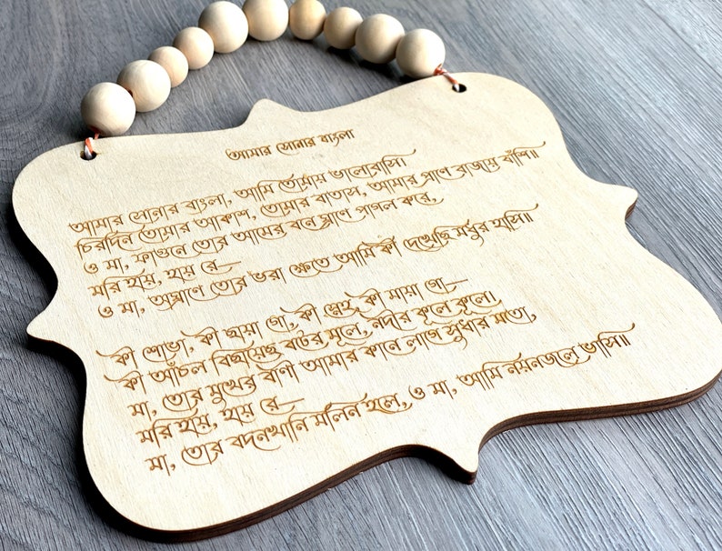 May include: A wooden plaque with a Bengali poem engraved on it. The poem is about love for one's homeland. The plaque is decorated with a string of wooden beads.