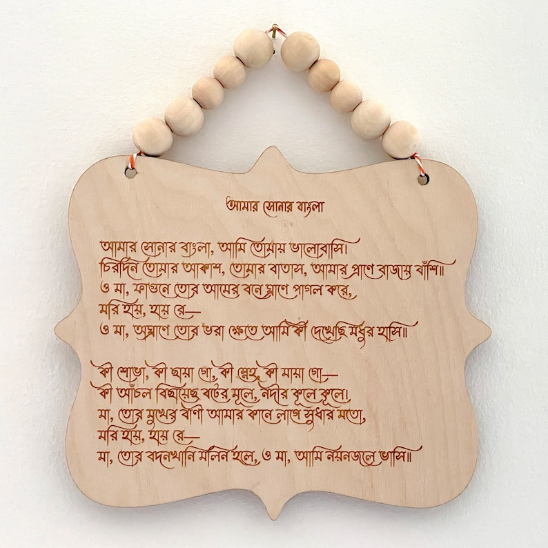 May include: A wooden plaque with a poem written in Bengali script. The poem is titled "Amar Sonar Bangla" and is about the beauty of Bangladesh. The plaque is hanging from a string of wooden beads.