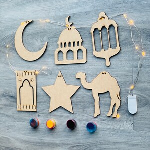 May include: A collection of wooden Ramadan decorations, including a crescent moon, mosque, lantern, star, camel, and prayer rug. The decorations are arranged with a string of fairy lights and four colorful, round magnets.