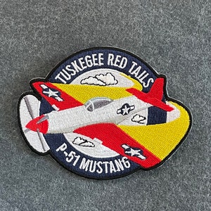 May include: A red, white, and blue embroidered patch with a cartoon P-51 Mustang airplane. The patch is circular and has the text "Tuskegee Red Tails P-51 Mustang" around the edge.