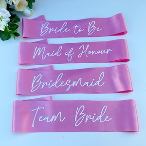 May include: Four pink satin sashes with white text. The sashes read "Bride to Be", "Maid of Honour", "Bridesmaid", and "Team Bride".