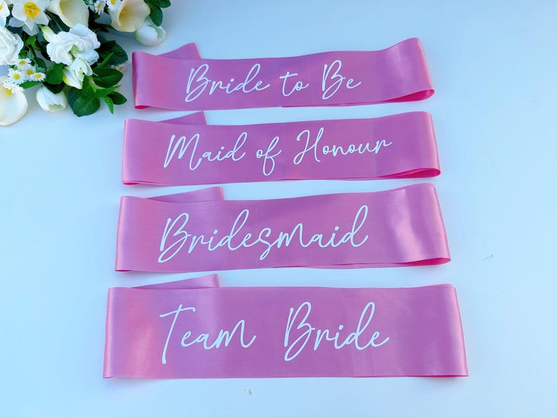 May include: Four pink satin sashes with white text. The sashes read "Bride to Be", "Maid of Honour", "Bridesmaid", and "Team Bride".