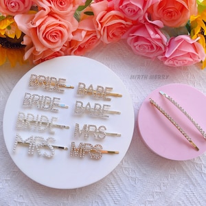 May include: A set of rhinestone hair clips with the words "Bride", "Babe", and "Mrs" on them. The clips are on a white background with a pink background behind them.