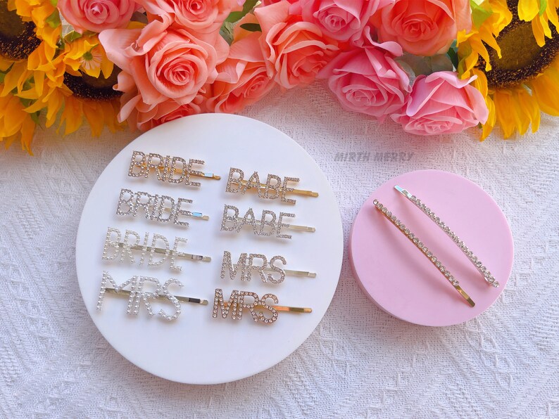 May include: A set of rhinestone hair clips with the words "Bride", "Babe", and "Mrs" on them. The clips are on a white background with a pink background behind them.