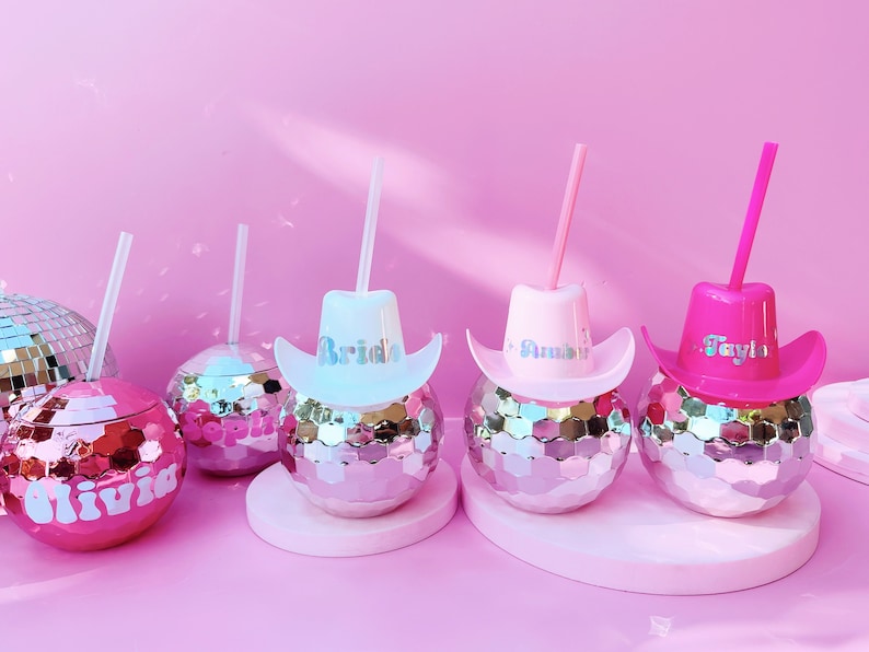 May include: Pink disco ball tumblers with cowboy hat lids and straws. The hats are in pink, white, and fuchsia, with names like "Bride", "Amber", and "Taylor". The tumblers are on a pink surface.