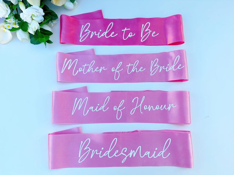 May include: Four pink satin sashes with white script. The sashes read "Bride to Be", "Mother of the Bride", "Maid of Honour", and "Bridesmaid".