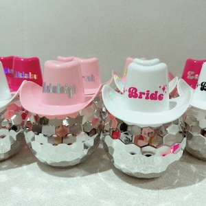 May include: Several disco ball-shaped objects, each adorned with a miniature cowboy hat. The hats are either white or pink, with the word "Bride" or a name printed on them. The disco balls have a mirrored, geometric design.