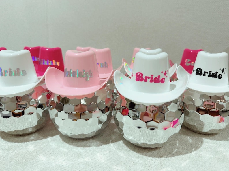 May include: Several disco ball-shaped objects, each adorned with a miniature cowboy hat. The hats are either white or pink, with the word "Bride" or a name printed on them. The disco balls have a mirrored, geometric design.