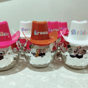 May include: A group of disco ball-shaped ornaments, each adorned with a small cowboy hat. The hats are in shades of pink, orange, and white, with holographic text displaying names and the words "Groom" and "Bride".