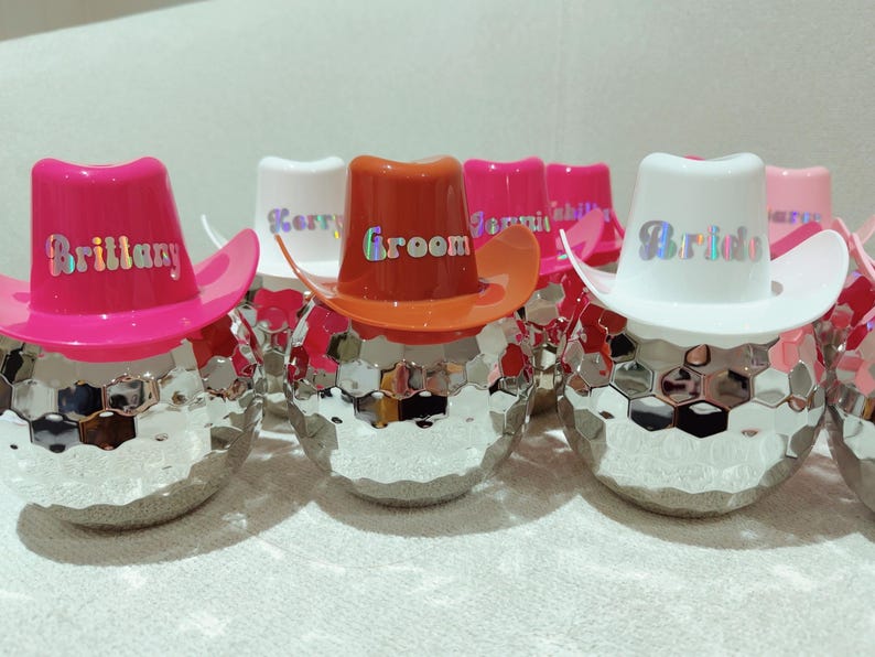 May include: A group of disco ball-shaped ornaments, each adorned with a small cowboy hat. The hats are in shades of pink, orange, and white, with holographic text displaying names and the words "Groom" and "Bride".