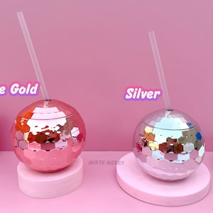 May include: Two disco ball-shaped tumblers, one rose gold and one silver, each with a clear straw. The tumblers sit on pink pedestals against a pink backdrop. Text on the image reads "Rose Gold" and "Silver". The tumblers are from "Mirth Merry".