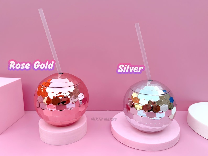 May include: Two disco ball-shaped tumblers, one rose gold and one silver, each with a clear straw. The tumblers sit on pink pedestals against a pink backdrop. Text on the image reads "Rose Gold" and "Silver". The tumblers are from "Mirth Merry".