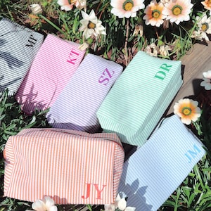 May include: Six striped fabric pouches with different coloured stripes and personalised initials. The initials are embroidered in different colours on each pouch. The pouches are arranged on a wooden surface with flowers in the background.