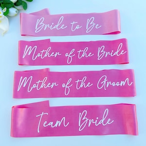 May include: Four pink satin sashes with white text. The sashes read "Bride to Be", "Mother of the Bride", "Mother of the Groom", and "Team Bride".