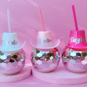 May include: Three disco ball tumblers with cowboy hat lids and straws. The hats are white, pink, and fuchsia, each with a name in holographic lettering: "Bride", "Amber", and "Taylor". The tumblers sit on a pink surface against a pink backdrop.