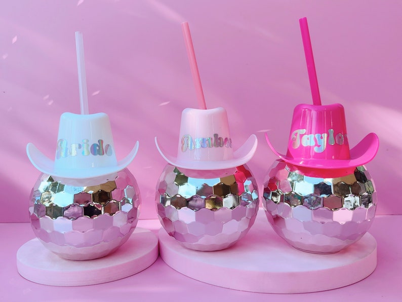 May include: Three disco ball tumblers with cowboy hat lids and straws. The hats are white, pink, and fuchsia, each with a name in holographic lettering: "Bride", "Amber", and "Taylor". The tumblers sit on a pink surface against a pink backdrop.
