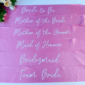 May include: Six pink satin sashes with white text. The sashes read "Bride to Be", "Mother of the Bride", "Mother of the Groom", "Maid of Honour", "Bridesmaid", and "Team Bride".