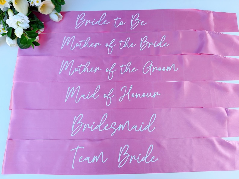 May include: Six pink satin sashes with white text. The sashes read "Bride to Be", "Mother of the Bride", "Mother of the Groom", "Maid of Honour", "Bridesmaid", and "Team Bride".