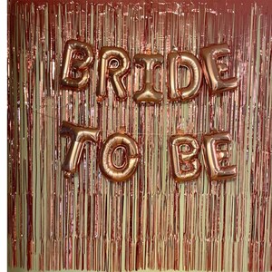 May include: Rose gold foil fringe backdrop with rose gold foil letter balloons spelling "Bride to Be".
