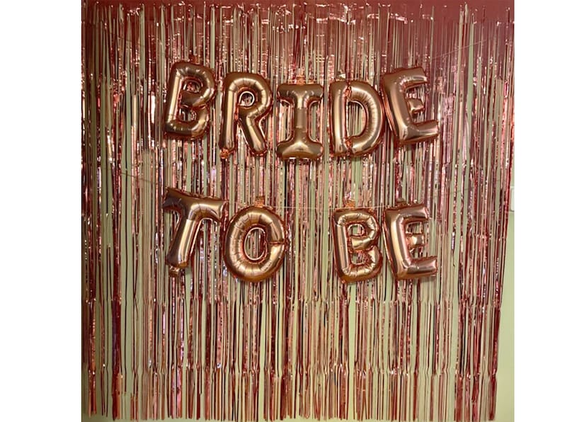 May include: Rose gold foil fringe backdrop with rose gold foil letter balloons spelling "Bride to Be".