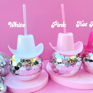 May include: Three disco ball-shaped tumblers with cowboy hat lids and straws in white, pink, and hot pink. The words "White", "Pink", and "Hot Pink" are written above each tumbler. The background is a solid pink colour.