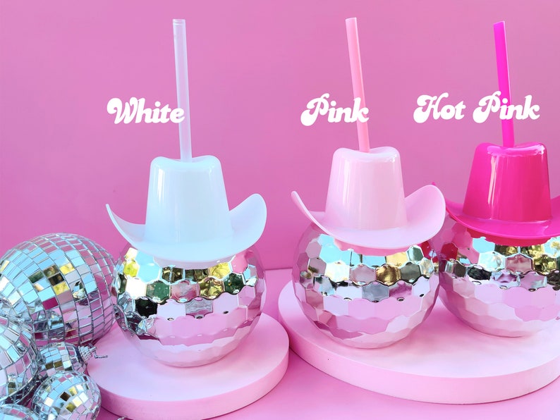 May include: Three disco ball-shaped tumblers with cowboy hat lids and straws in white, pink, and hot pink. The words "White", "Pink", and "Hot Pink" are written above each tumbler. The background is a solid pink colour.