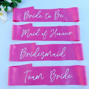 May include: Four pink satin sashes with white text. The sashes read "Bride to Be", "Maid of Honour", "Bridesmaid", and "Team Bride".