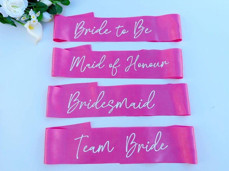 May include: Four pink satin sashes with white text. The sashes read "Bride to Be", "Maid of Honour", "Bridesmaid", and "Team Bride".