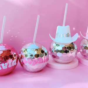 May include: Pink disco ball-shaped tumblers with white straws. Two tumblers have names in white script, and two have miniature cowboy hats with names. The tumblers sit on a pink surface against a pink background.