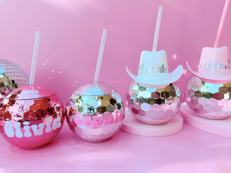 May include: Pink disco ball-shaped tumblers with white straws. Two tumblers have names in white script, and two have miniature cowboy hats with names. The tumblers sit on a pink surface against a pink background.