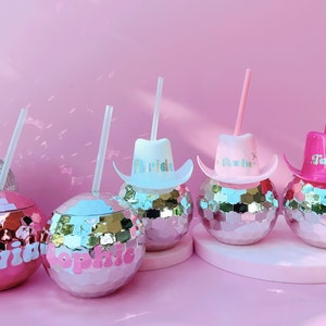 May include: Pink disco ball tumblers with straws, personalised with names and cowboy hat lids. The tumblers have a mirrored surface and are set against a pink backdrop, ideal for a party or celebration.