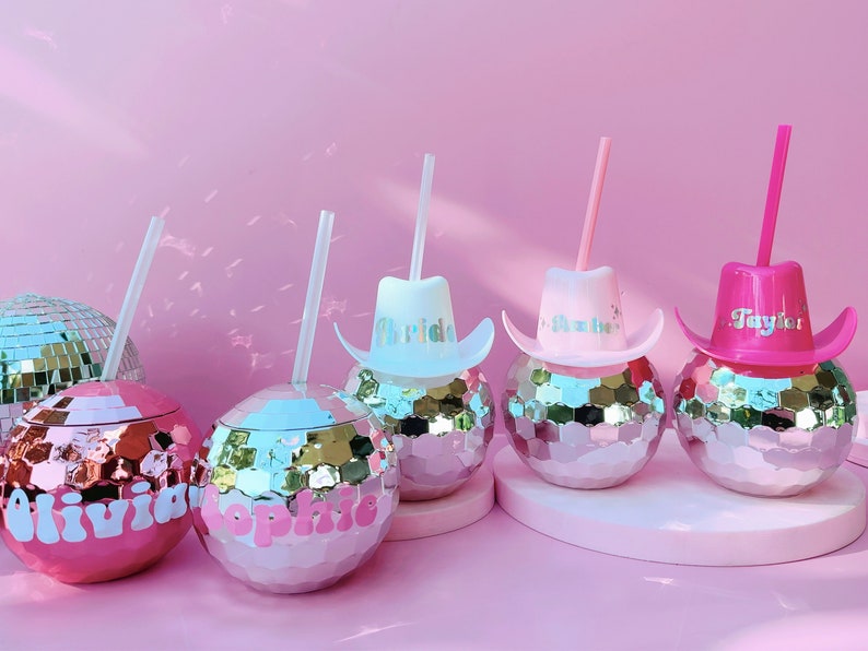 May include: Pink disco ball tumblers with straws, personalised with names and cowboy hat lids. The tumblers have a mirrored surface and are set against a pink backdrop, ideal for a party or celebration.