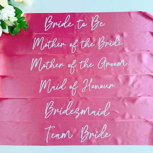 May include: Six pink satin sashes with white text. The sashes read "Bride to Be", "Mother of the Bride", "Mother of the Groom", "Maid of Honour", "Bridesmaid", and "Team Bride".