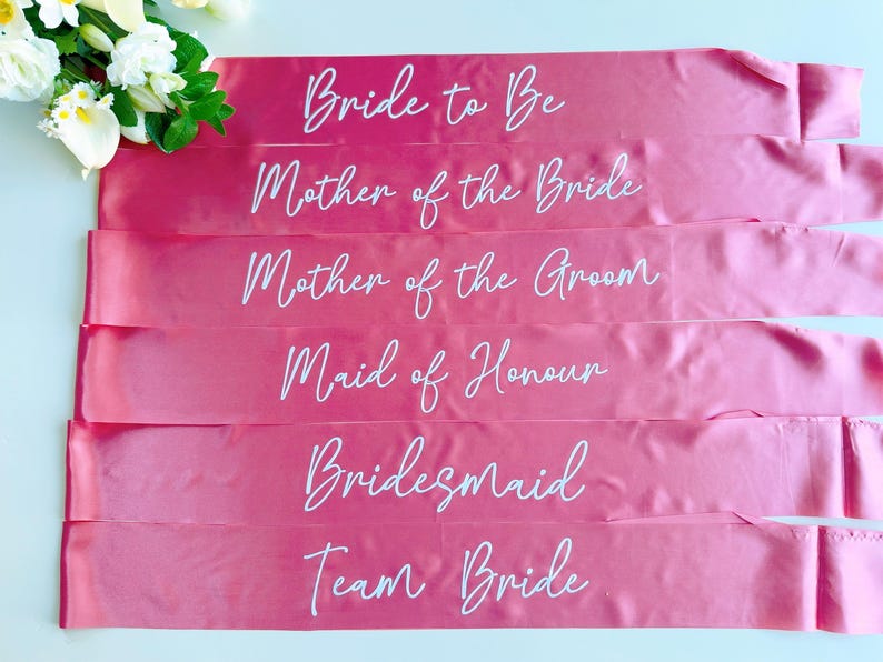 May include: Six pink satin sashes with white text. The sashes read "Bride to Be", "Mother of the Bride", "Mother of the Groom", "Maid of Honour", "Bridesmaid", and "Team Bride".