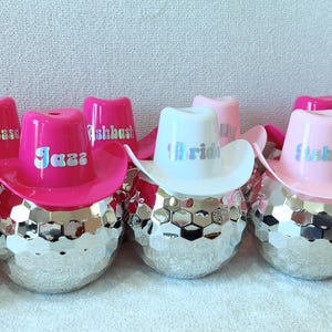 May include: A selection of decorative items, each with a disco ball base and a miniature cowboy hat. The hats are in pink and white, with names such as "Jazza" and "Ashley". The "Bride" hat is white.