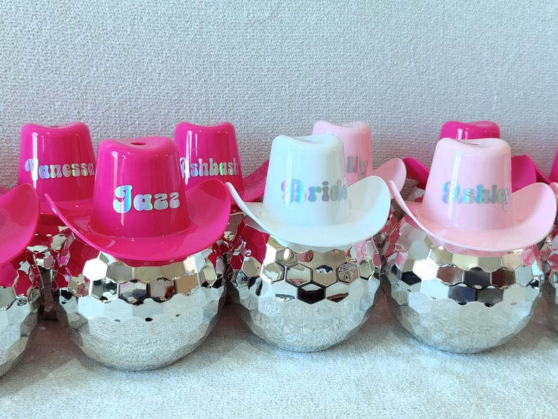 May include: A selection of decorative items, each with a disco ball base and a miniature cowboy hat. The hats are in pink and white, with names such as "Jazza" and "Ashley". The "Bride" hat is white.