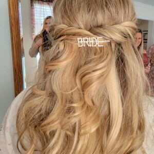 May include: A woman with long, blonde hair is wearing a white dress and has a hair clip with the word "BRIDE" in rhinestones in her hair.