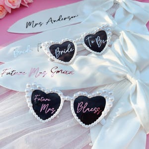 May include: Three pairs of heart-shaped sunglasses with pearl accents. The sunglasses have black lenses and white text that reads "Bride", "To Be", and "Future Mrs. Bless".