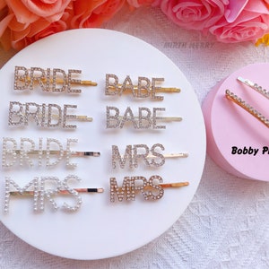 May include: Assortment of rhinestone hair clips with the words "Bride", "Babe", and "Mrs" on a white background.  Two bobby pins with rhinestones are on a pink background.