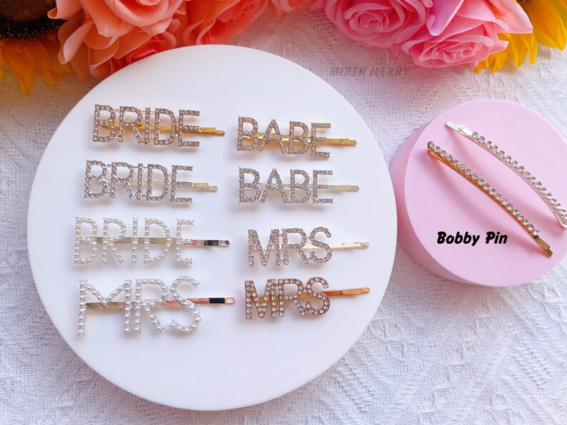 May include: Assortment of rhinestone hair clips with the words "Bride", "Babe", and "Mrs" on a white background.  Two bobby pins with rhinestones are on a pink background.