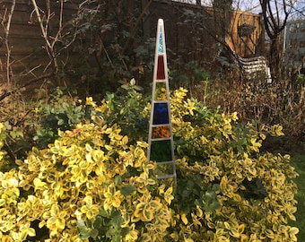 May include: A colorful stained glass garden stake with red, orange, yellow, green, blue, and clear glass pieces. The stake is in a garden setting with green and yellow foliage.