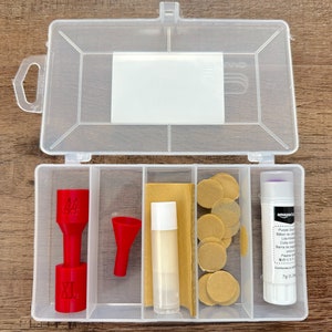 May include: A clear plastic organizer box with multiple compartments. The box contains red plastic pieces, a lip balm, sandpaper, round tan stickers, and a glue stick with text. The box is open, revealing the contents.