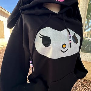May include: Black hooded sweatshirt featuring a white and black cartoon face design on the front. The hood has pink bunny ears and a pink skull graphic. The drawstring has decorative beads.