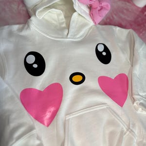 Puede incluir: Sudadera con capucha blanca con un lazo rosa en la capucha y una cara de dibujos animados con corazones rosas y ojos negros en la parte delantera.