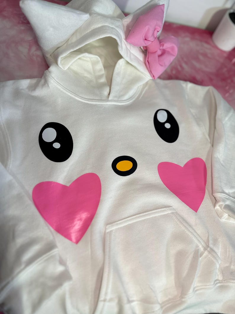 Puede incluir: Sudadera con capucha blanca con un lazo rosa en la capucha y una cara de dibujos animados con corazones rosas y ojos negros en la parte delantera.